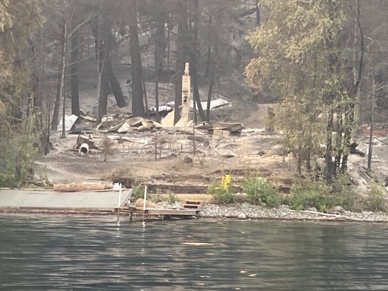 The ruins of a cabin on a lake are barely standing amidst scorched trees.