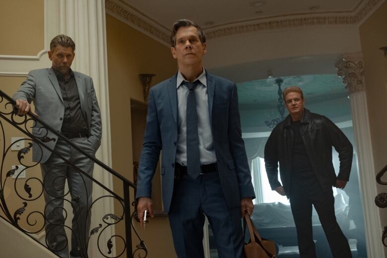 3 men stand in a hallway.  Actor Kevin Bacon is in the middle. 