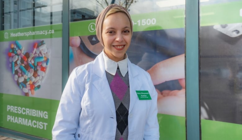 Heba Elbayoyumi wears a white lab coat as she stands outside her pharmacy. A green sign with pills gathered in the shape of a heart can be seen behind her.