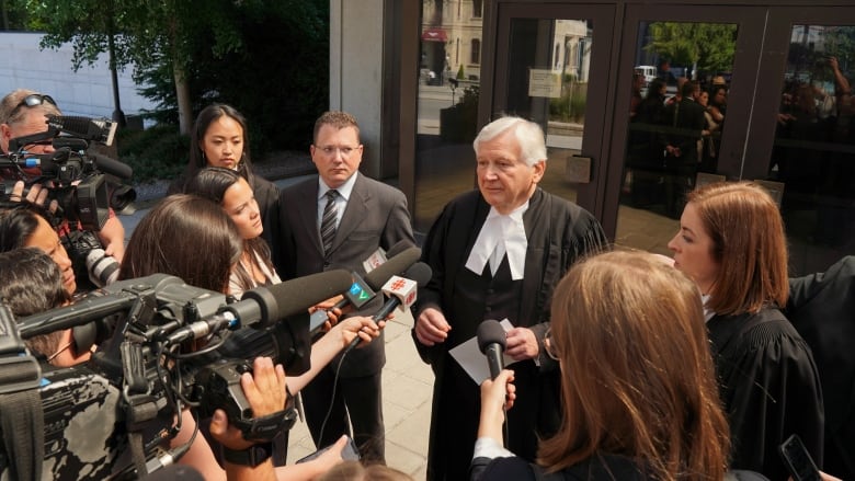 A lawyer talks to reporters outside a courthouse in summer. His client is beside him.