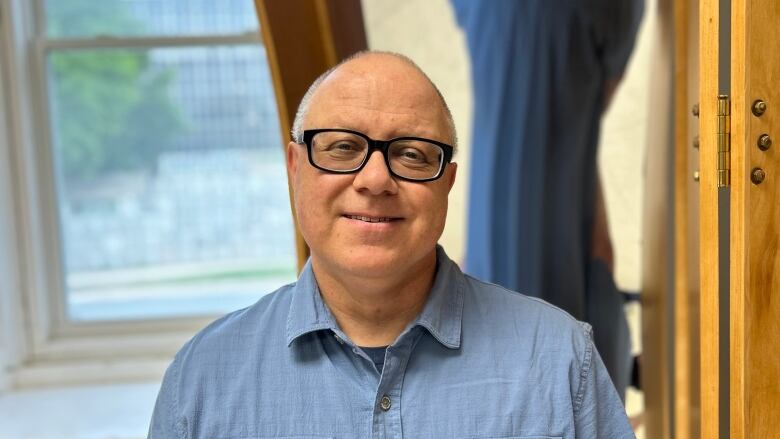 A man in about his 50s with short gray hair and wearing black rimmed eye glasses and a short-sleeved medium blue shirt smiles towards the camera for an upper body portrait. In the background on the right is a curved mirror showing a distorted reflection of his backside. In the back on the left is a window of a tree and part of an office building.