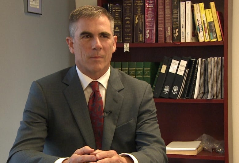 A man in a grey suit and dark red tie sits in front of bookshelves lined with books and binders.