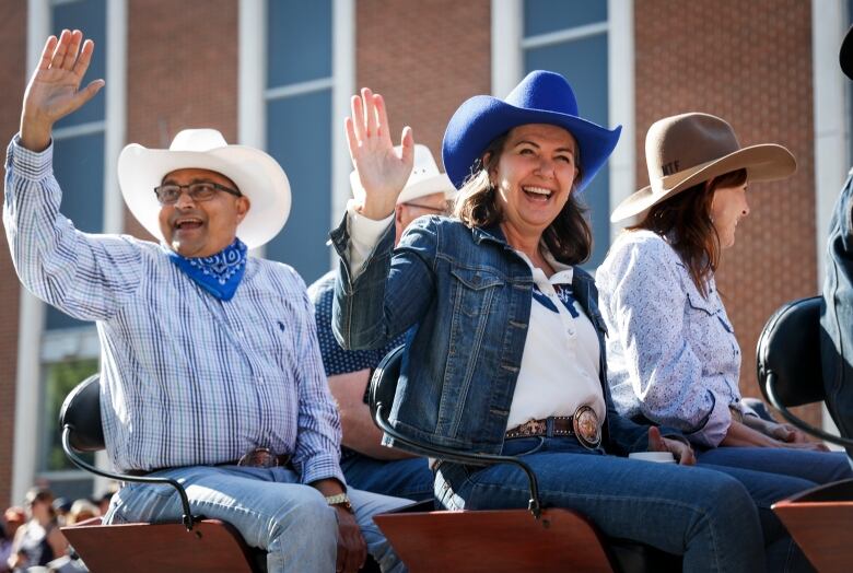 Three people riding in the back of a wagon. In the middle, Premier Danielle Smith, wearing a blue cowboy hat, smiles and waves to the crowd.