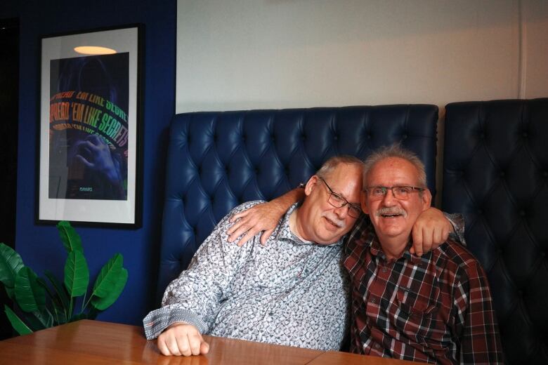 Two men who look to be in their 60s pose for a photo, with their arms around each other. The one on the left wears a patterned shirt, while the one on the right wheres a plaid shirt. Both are wearing glasses. They're sitting on a blue booth, next to a framed poster that says: Spread 'em like secrets.