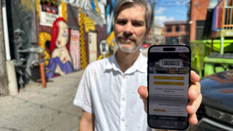 Parallel42 owner Doug Sartori holds his cellphone connected to the MotorCityMurals.tours app for self-guiding tours of the murals in Windsor and Detroit while standing in the WIFF Alley mural area.