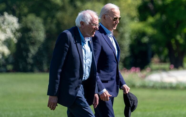 Two white-haired men, wearing glasses and navy blazers, walk in a park.