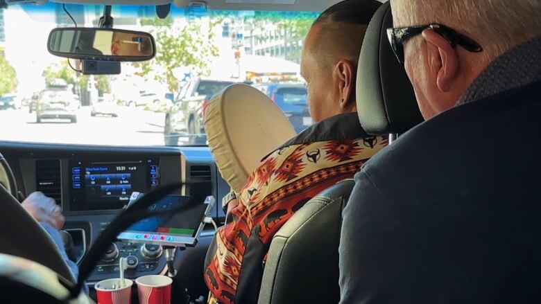 A man sits in the front of a cab, a passenger behind him, you can see the backs of their heads. 