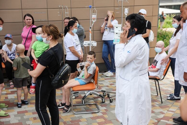 Hospital workers and civilians stand around outdoors, with some children seen with IV's.