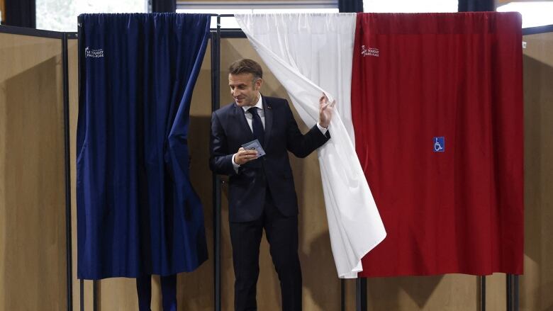 A man in a dark suit appears from behind a French flag draped over a voting booth.