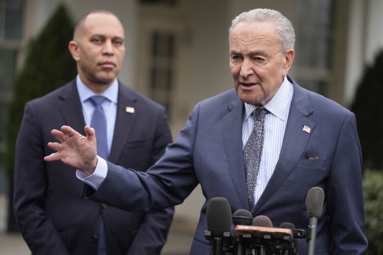 Two clean-shaven men in suit and tie are shown outdoors, with the older-looking of the two speaking and gesturing as the other looks on.