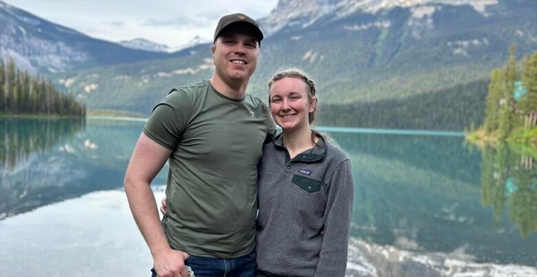 Man wearing navy green shirt and cap stands besides a woman wearing a gray sweater. Both of them stand in front of lake and mountain backdrop, smiling at the camera.