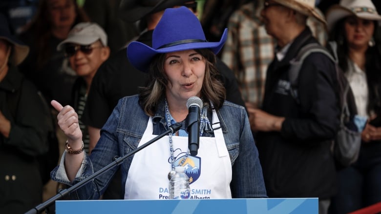 Alberta Premier Danielle Smith speaks at a podium at her pancake breakfast, wearing a blue cowboy hat, apron and jean jacket. The podium signage says Alberta.