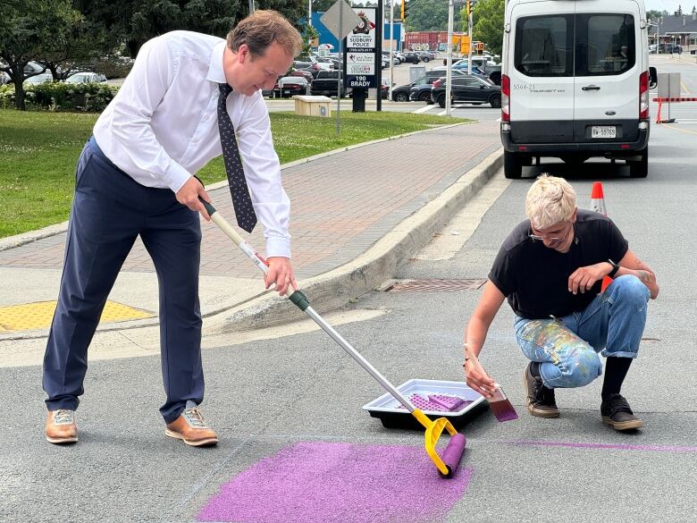 A person wearing a tie paints the ground with another person sitting on the floor painting.