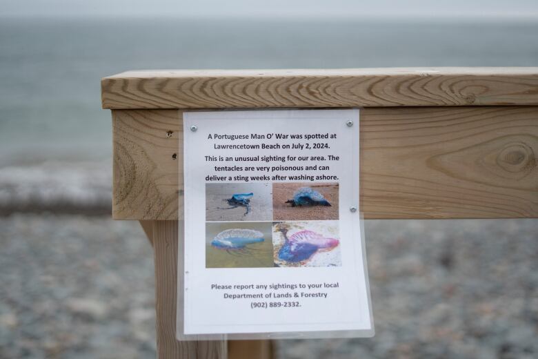 a laminated sign hangs on a wooden rail near a beach.