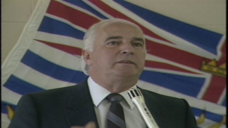 A man in a suit in front of the B.C. flag.