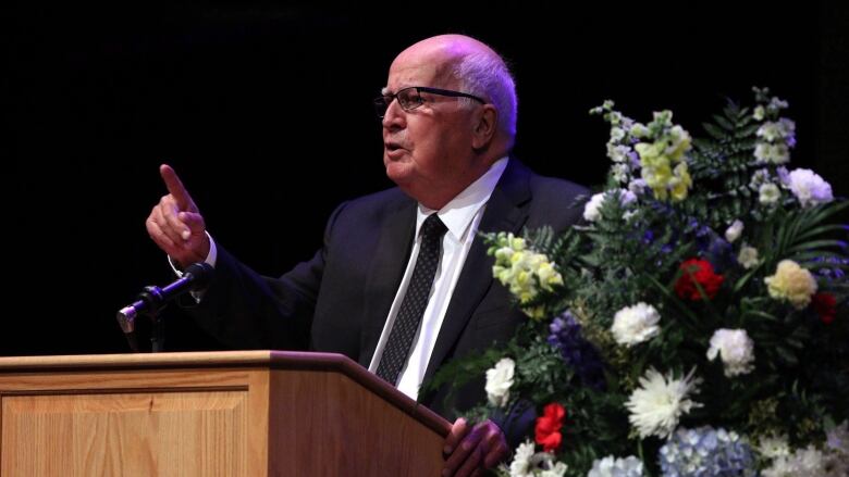 A man waves his finger as he speaks at a podium, with flowers visible to his left.