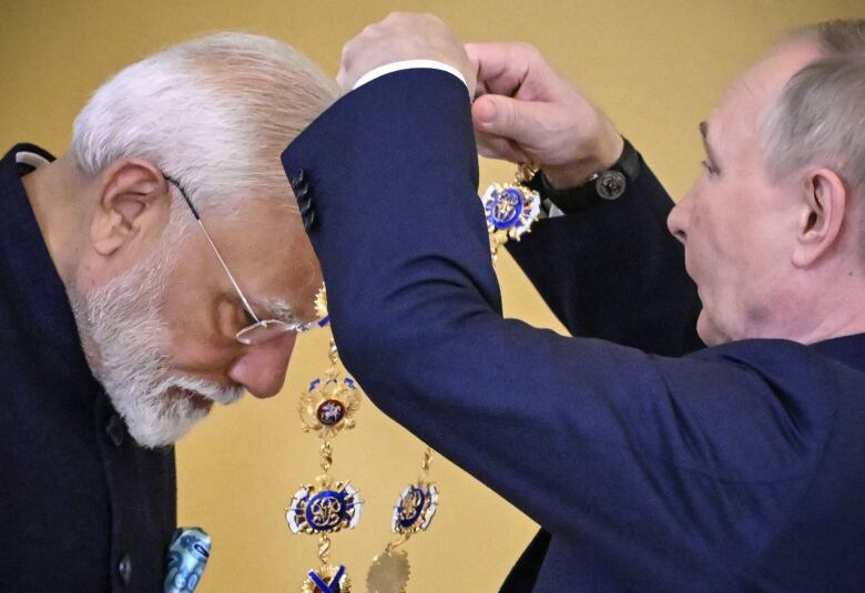 A white-bearded man in glasses leans over to accept a large necklace of medallions about to be place around his neck by a cleanshaven man wearing a suit jacket.