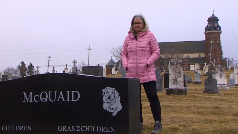 A woman in a pink jacket is shown next to a black gravestone, with a church in the background.