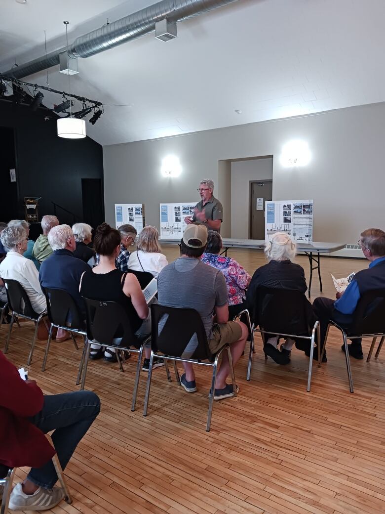 A old man with a grey collared shirt talking to a small group of 20 people who are sitting in chairs 