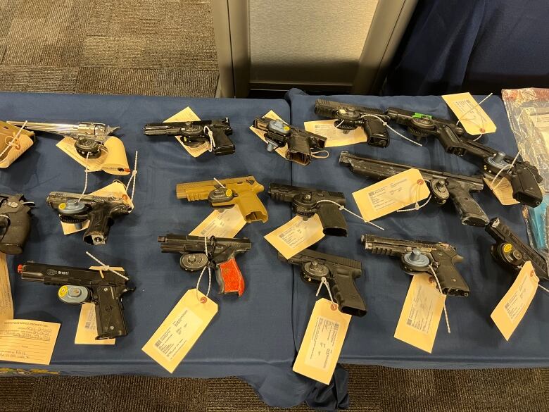 Handguns in various sizes and colours, with paper tags attached to each one, are laid out on a dark blue tablecloth on a table.