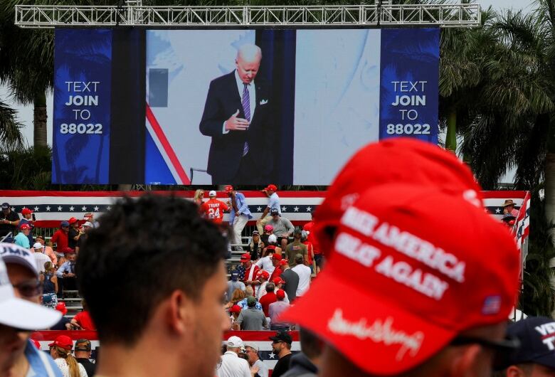 A red MAGA cap, with a screen in the background showing an image of Biden.