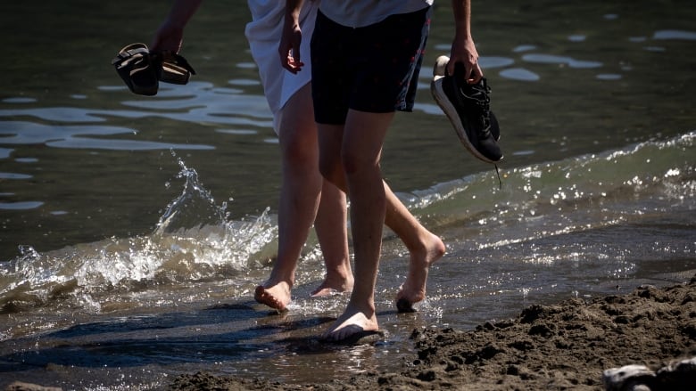 Two people walk barefoot along water.