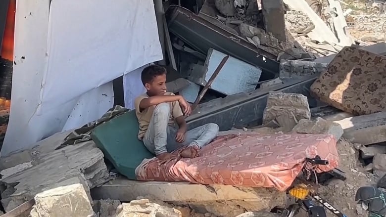 A boy sits on  a tattered mattress in a  pile of rubble