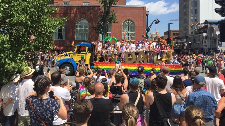 A crowd of people watch as a parade float decorated with a large rainbow on the side and with people on it goes by.