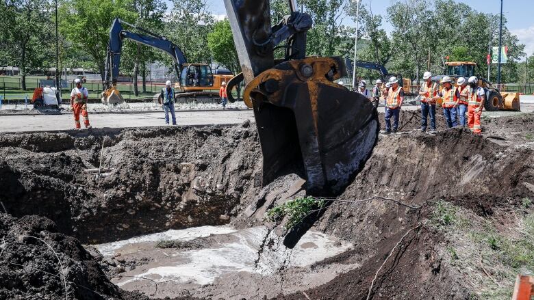 construction workers stand around a large hole filled with muddy water as a large piece of construction equipment digs into the hole.