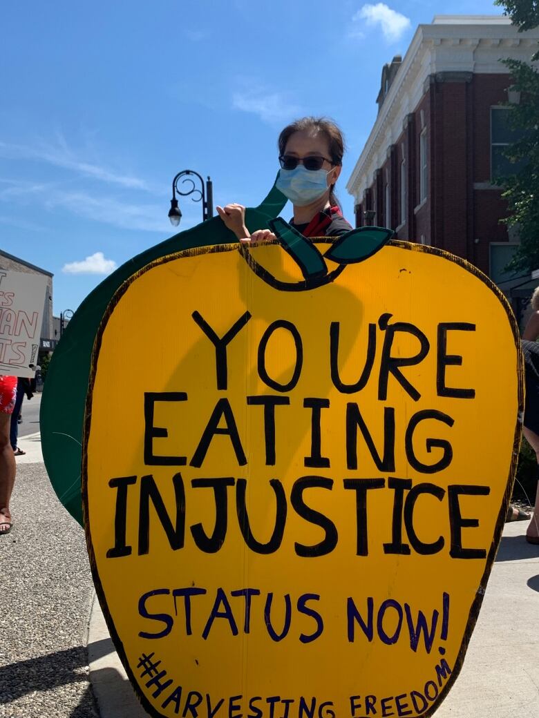 Demonstrator in mask and sunglasses holds sign in the shape of an apple that reads: 