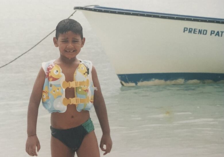 A young boy with a gap in his teeth and wearing a swimsuit and lifejacket stands in front of a body of water, a ship behind him.