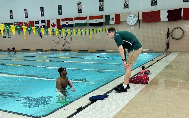 A man in a swim cap treads water in a pool while a man in a green shirt and khaki shorts leans over to speak to him from the side of the pool. 