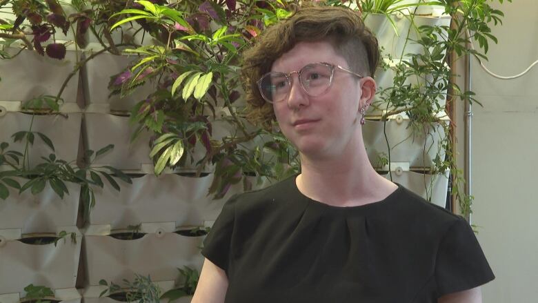 A person with short hair shaved on one side and wearing glasses. They are wearing a black t-shirt and standing in front of a wall of plants.