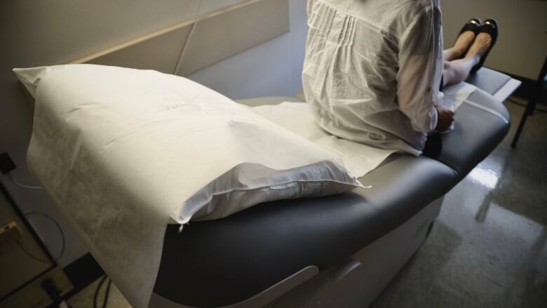 A woman, whose face is not visible, sits on an exam table with table paper draped over it