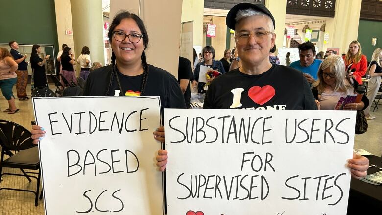 Two people hold signs in support of supervised consumption sites and substance users.