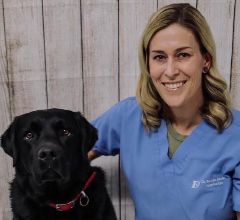 A blonde woman in blue scrubs posing with a black lab.