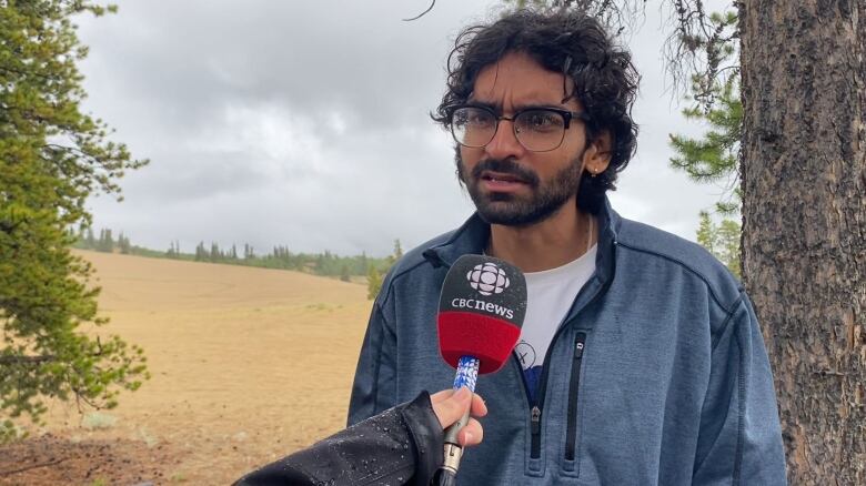 A man standing near some trees and sand speaks into a CBC microphone.