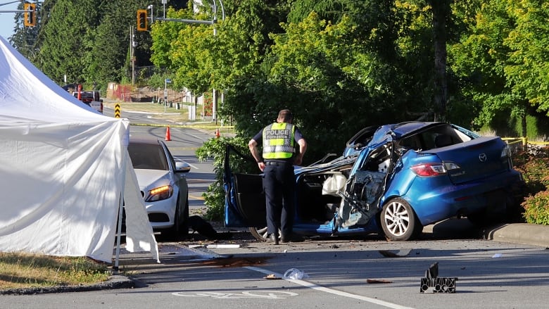 A police officer looks at a mangled sedan next to a white tent on a city street.