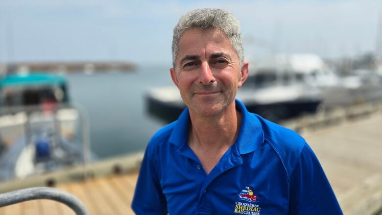 A man with short grey hair and wearing a blue shirt stands on a dock. Boats float behind him.