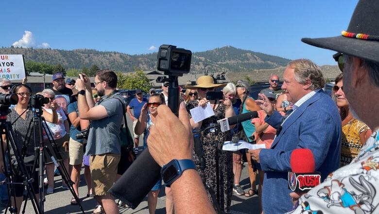 A gathering of people, including reporters with mics and cameras, in an outdoor environment with hills in the background.