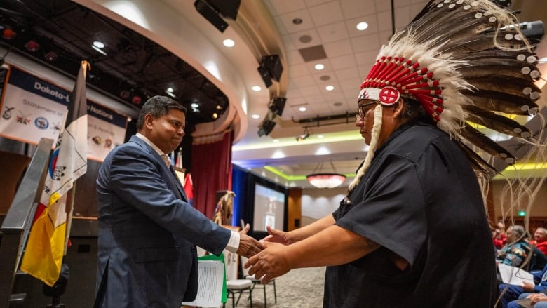 A man in a business suit shakes hands with a man wearing a traditional Indigenous headdress.