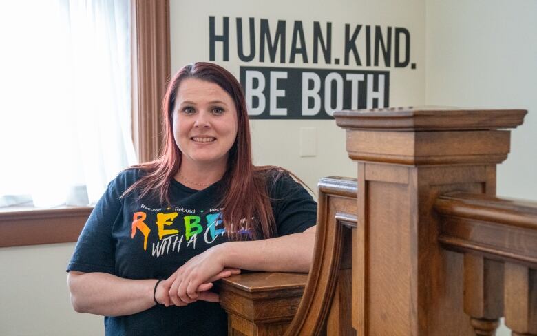 A person is seen leaning at the top of a wooden bannister, smiling. Behind them is a sign on the wall that says, 