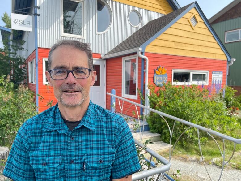 A man in glasses and a blue plaid shirt smiles for a photo outdoors in front of a brightly coloured house.