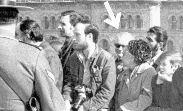 An old black and white photograph shows several individuals gathered in a crowd in an outdoor setting, with an arrow added to point to a man wearing dark sunglasses.