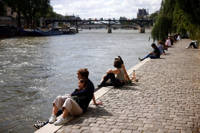 People sit along the Seine river banks. A couple in the foreground leans on each other.