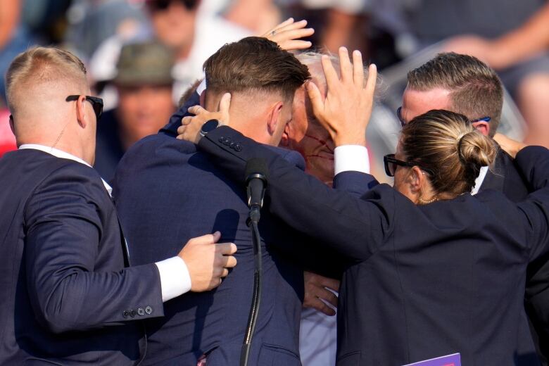 People in dark suit jackets are shown gathered and huddled in close proximity in an outdoor setting.