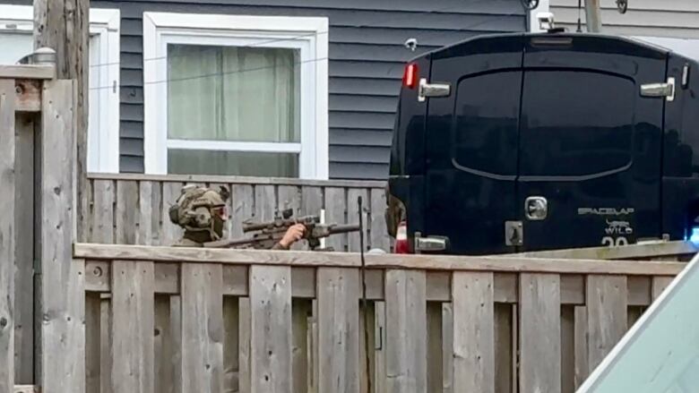 A police tactical officer where camo and a helmet hold up a rifle behind a wood fence.