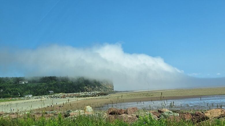 Fog rolls through a blue sky over a sandy shore.