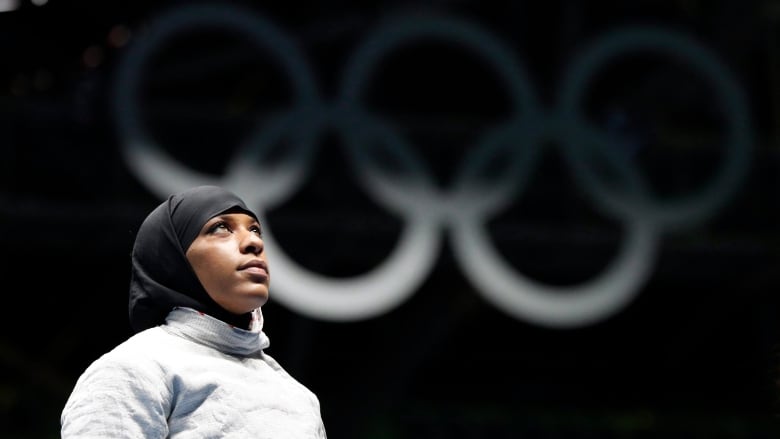 A woman in a hijab stands in front of the Olympic rings.