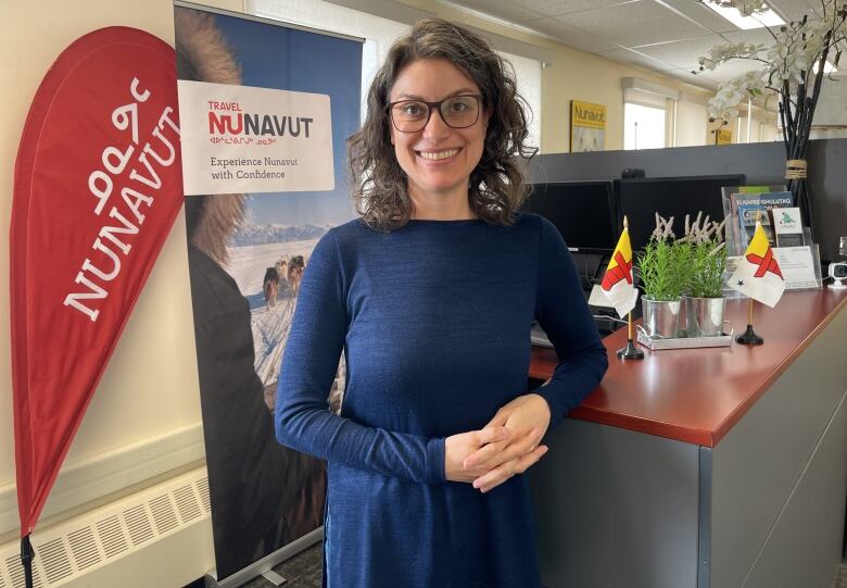 A woman with brown hair wearing a blue dress and glasses standing beside a desk with two flags and a plant are resting on it